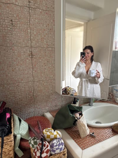 The scene depicts an interior bathroom within the Anavissos Municipal Unit, Greece. A female individual is reflected in a large rectangular mirror positioned above a white ceramic sink with a chrome single-lever faucet. She has dark, damp hair and wears a white waffle-knit bathrobe tied at the waist, revealing a gold-colored necklace. She is holding a smartphone with a dark case in her left hand, capturing her reflection, and a white mug with a green cactus graphic in her right hand. The wall to the left of the mirror and sink is covered in reddish-brown and off-white mosaic tiles with dark, cracked grout lines. The countertop around the sink is similarly tiled. On the right side of the sink, a clear bottle labeled "ADROSORPT PLUS," a dark pump bottle, and a wooden hairbrush are visible. On the left side of the sink, a dark green zippered pouch contains makeup brushes, next to a smaller off-white cloth bag also holding brushes, and a silver hair clip. Below the counter, on a tiled surface matching the wall, two woven wicker baskets are present. The left basket contains a black and pink hairdryer attachment and additional hair tools. The right basket holds four rolled-up, checkerboard-patterned hand towels in yellow/white, green/white, purple/white, and reddish-brown/white. A light green woven bag, a circular reddish-brown woven mat, and a rose gold-colored hair styling tool are also visible in the foreground.