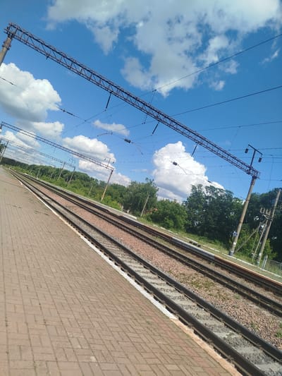 The image shows a railway station platform and tracks in Chervonyi Step, Ukraine. The platform is paved with brick pavers. Multiple sets of railway tracks run parallel to the platform. Overhead power lines, supported by metal structures and concrete poles, stretch across the scene, indicating an electrified railway line. The background features a line of trees and a blue sky with scattered white clouds. No people or vehicles are visible. The image is taken from a low angle, looking down the tracks.