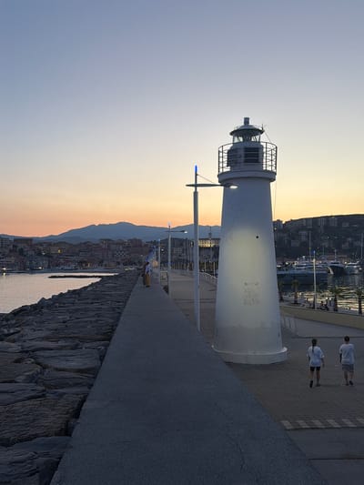 The image shows a lighthouse on a pier at dusk, with a coastal town and mountains in the background. Two people, likely teenagers, are walking away from the camera on the paved walkway. The lighthouse is white and cylindrical, with a metal cage around its lantern room. A single blue light is visible on a pole next to the lighthouse. The sky is a gradient of pale blue and orange, indicating sunset or sunrise. The town in the background is illuminated by lights, suggesting it is either late evening or early morning. The sea is calm and reflects the lights of the town. The pier is made of stone and concrete. The overall mood is peaceful and serene. The location appears to be Imperia, Italy, given the context.
