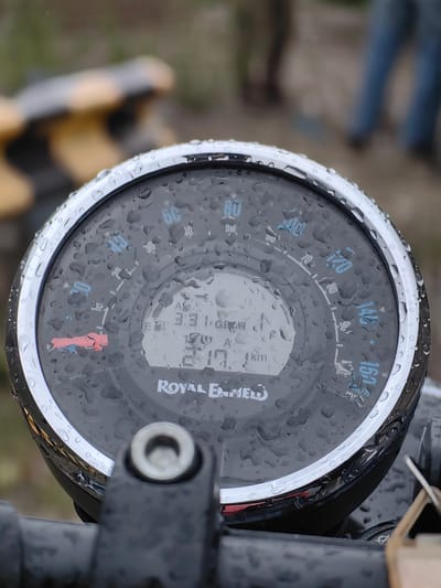 The image is a close-up shot of a motorcycle's speedometer, which is wet with raindrops. The speedometer has a chrome bezel and a black face with white markings for speed in km/h, ranging from 0 to 160. A digital display in the center shows "3:31" and "13". The "Royal Enfield" logo is visible at the bottom of the speedometer face. A red indicator light is illuminated on the left side of the speedometer. In the blurred background, there are indications of an outdoor environment with people in the distance, suggesting it might be a public area or a gathering. The overall impression is of a motorcycle parked outdoors during or after rainfall. The location of Chikka, India, is not discernible from the visual information in the image itself.