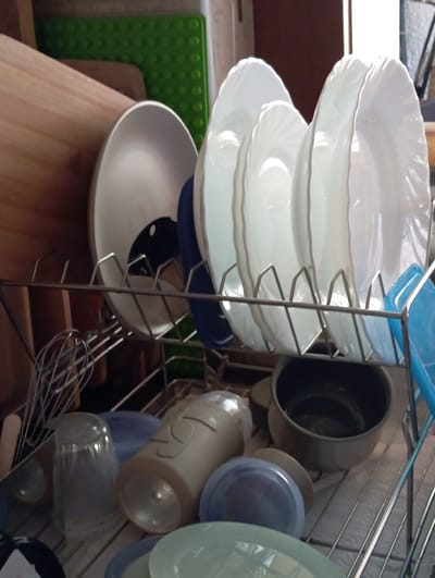A close-up, eye-level shot shows a chrome dish rack filled with various white plates and bowls, some with fluted edges. The plates are stacked neatly, with the largest at the bottom. Beneath the plates, a blue bowl and a large, dark pot are visible. To the right, a beige plastic container with embossed markings, a blue bowl, and a lighter blue-green bowl are also present. Below these, a clear plastic container and a whisk are positioned. In the background, a green textured mat is partially visible. The setting appears to be a kitchen in Baden, Austria, suggesting the dish rack is either in use or being stored. The lighting indicates it is daytime.