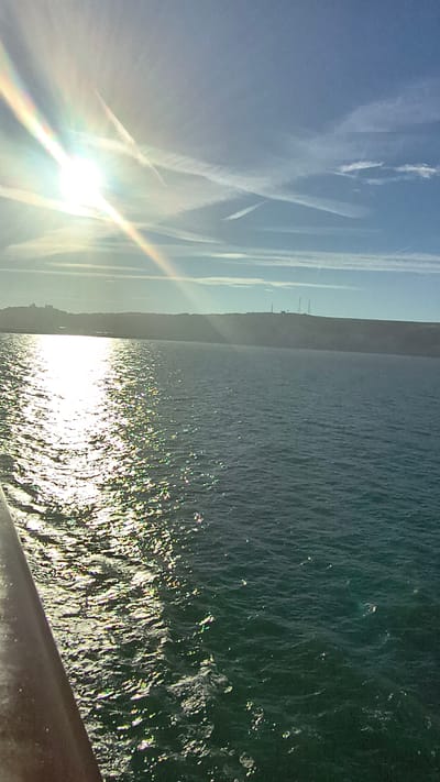 This media file depicts a scenic view of the sea and sky, likely captured from a high vantage point like a cliff or a bridge in Dover, United Kingdom. The image is oriented vertically, with the sky occupying the left half and the sea the right.
The sky is a clear, bright blue, with streaks of white contrails crisscrossing its expanse, suggesting recent airplane activity. The sun is visible as a brilliant, lens-flared orb, casting a warm, bright light across the sky and creating a rainbow-like halo.
The sea is a deep, dark blue-green, with gentle waves creating a textured surface. The sun's reflection glitters intensely on the water, adding a shimmering effect. In the distance, along the horizon where the sea meets the sky, a dark, elevated landmass is visible, likely the White Cliffs of Dover. The silhouette of what appear to be industrial structures, possibly smokestacks or cranes, can be discerned atop this landmass, hinting at the presence of human activity or infrastructure near the coastline.
The overall mood of the image is tranquil and expansive, evoking a sense of the vastness of nature. The bright sunlight suggests it is daytime, likely mid-morning or afternoon, given the angle of the sun. The weather appears clear and sunny. No people or specific objects are the primary focus, rather the natural elements of sea and sky are the main subjects, with the distant coastline serving as a contextual element. There is no visible text in the image.