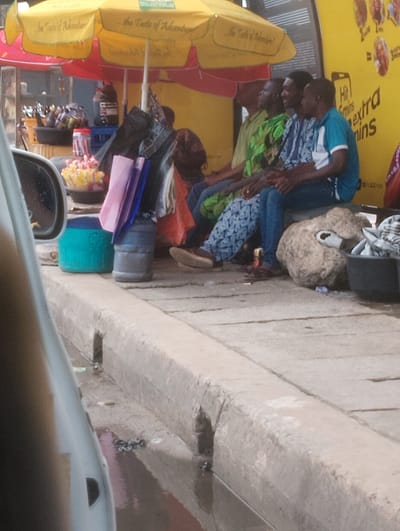 The image depicts a street scene in Oworonshoki, Nigeria, with a group of men sitting. Two yellow umbrellas, likely for shade, are visible. One has red trim and text that reads "... the Taste of Adventure!" The other umbrella is primarily yellow. In the background, a bright yellow wall with text in English suggests an advertisement or signage. The men are seated on a low, concrete structure, with some wearing t-shirts and others in patterned shirts and blue jeans.
In the foreground, a grey vehicle, possibly a car, is partially visible on the left. A concrete curb runs along the bottom of the image. Scattered items on the ground include a blue plastic container, a dark grey cylindrical object, and other miscellaneous objects, some appearing to be wrappers or debris. A large, rough, light brown rock is positioned near the seated individuals, with a white plastic bag protruding from it. A dark plastic basin containing what appears to be fabric or clothing is also present. The ground is paved with concrete slabs and shows signs of wetness and some discarded items. The overall environment appears to be an outdoor public space, likely a market or roadside area.