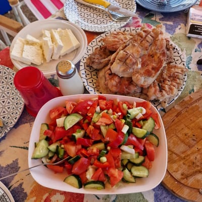 The image depicts a close-up view of a meal laid out on a table, possibly during an outdoor gathering in Gorni Dabnik, Bulgaria. In the foreground, a white bowl is filled with a fresh salad consisting of chopped tomatoes, cucumbers, red bell peppers, and green onions. A fork rests in the salad, suggesting it is ready to be eaten. To the left of the salad bowl, a red plastic cup is visible. Behind the salad and cup, on a plate with a geometric pattern, are several slices of white cheese. Further back, another plate displays grilled chicken. A salt shaker with a metallic top is positioned between the cheese and the grilled chicken. In the upper right corner, a pack of cigarettes is partially visible. The table is covered with a colorful tablecloth featuring floral and abstract patterns. The overall scene suggests a casual meal with a focus on fresh ingredients and grilled food.