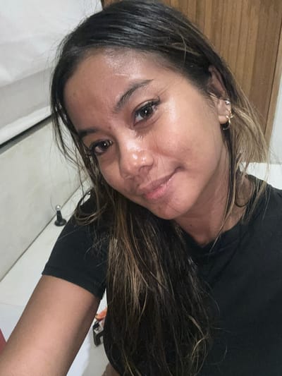 A close-up, rotated image depicts a woman with dark hair streaked with lighter brown highlights. She is wearing a black shirt. She has visible ear piercings on her left ear. Her skin appears shiny, possibly due to sweat. The background includes a wooden door and a white wall. No information to indicate this is in Tumbak Bayuh, Indonesia.