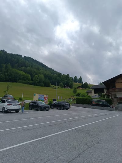 The image depicts a parking lot in Hallein, Austria, with a mountain landscape in the background under a cloudy sky. Several vehicles are parked in the lot, including a white SUV, a black SUV, and another dark-colored car. A woman is walking in the parking lot carrying a child. In the background, a green, tree-covered mountain slopes upwards. Several buildings, characteristic of Austrian architecture with wooden accents, are visible on the hillside. A ski lift or gondola system can be seen ascending the mountain. The overall atmosphere is overcast, suggesting it may be a cool or rainy day. A black van with "DACHSFEZI" and "Spenglerei & Bedachungen" written on its side is parked near a building. A small playground structure is also visible on the left side of the image.