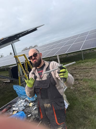 The image depicts an outdoor solar farm construction site in Baldurgan Hill, Ireland, under an overcast sky. The ground consists of green grass with visible muddy patches. In the background, multiple rows of dark grey solar panels are installed on metal support structures, extending into the distance. A bald adult male with a dark beard, wearing sunglasses, a light grey hooded jacket, dark grey overalls with orange accents, and yellow-green work gloves, is positioned centrally, facing the camera. He displays a pouting expression. His left hand forms a "rock on" gesture, while his right hand holds a silver, T-shaped metal tool or component. In the immediate foreground, a flat surface is covered with numerous small silver metal clips, nuts, and bolts, alongside a red-handled power tool and blue plastic bags containing debris. A yellow step ladder is partially visible behind the individual.