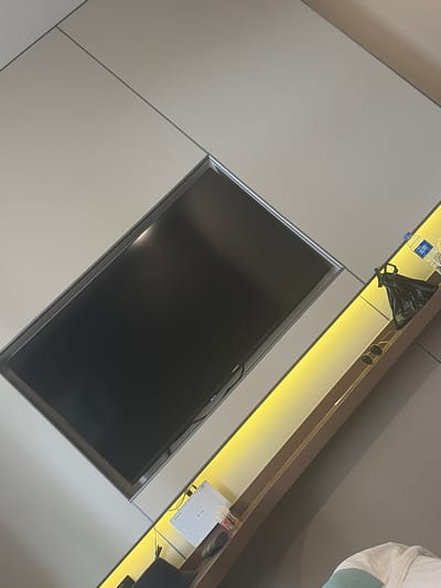 The image displays a modern living room interior, likely in Aihui, China. A large, dark flat-screen television is embedded in a light-colored wall panel. Below the television, a long shelf with a warm yellow LED backlight showcases several items. These include two pairs of sunglasses, a black trash bag, and two plastic water bottles, one with a blue cap and Chinese writing on the label. To the left of the shelf, a white electronic device, possibly a router or modem, is connected to power. The overall aesthetic is minimalist and contemporary, with clean lines and subtle lighting. The room appears to be well-lit, with natural light perhaps entering from an unseen source.