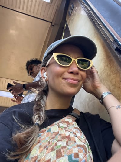 The image is a selfie taken by a woman wearing a grey baseball cap, yellow sunglasses, and a black t-shirt. She has a braid of hair extending over her shoulder and is wearing white wireless earbuds. A silver watch is visible on her right wrist, along with a bracelet with a charm. Behind her, a man wearing glasses and a checkered shirt is holding a black smartphone. The background appears to be the interior of a train carriage, with worn beige walls and vertical ribbed panels. A sign with the number "2" is visible on the wall behind the man. The overall lighting suggests it is daytime. This scene is consistent with being inside a train carriage of the Sri Lanka Railways, likely operating on a route that passes through Kalutara, a city known for its coastal train line.
