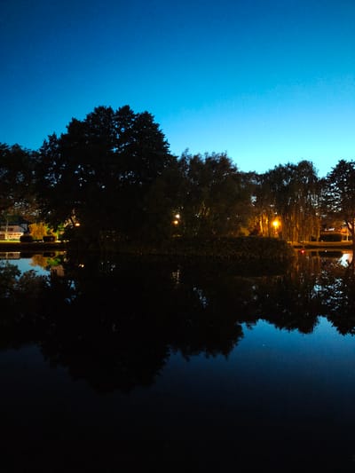 The image depicts a serene twilight scene at the Rumia Reservoir in Rumia, Poland. Silhouetted trees and foliage line the banks of the dark, still water, their reflections creating a mirror image on the surface. In the distance, artificial lights from buildings and streetlamps cast a warm glow, contrasting with the deep blue of the twilight sky. The overall atmosphere is tranquil and peaceful, with a hint of urban life subtly present.