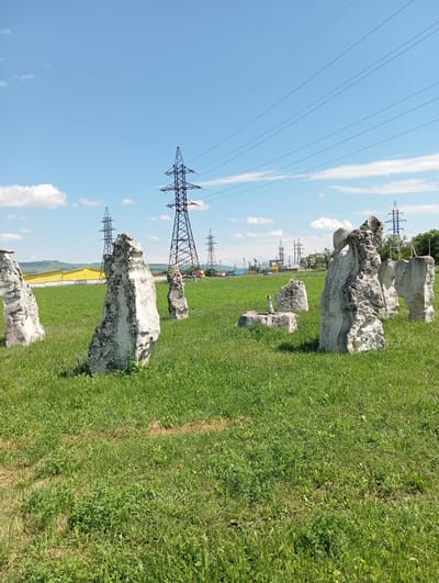 The image depicts an outdoor scene with several large, weathered stone formations standing in a grassy field under a bright blue sky with scattered white clouds. In the background, several tall, metal power line towers and high-voltage wires are visible, indicating the presence of significant electrical infrastructure. A yellow building with a white fence is also visible in the mid-ground. The overall environment appears to be rural or semi-rural, with rolling hills in the far distance. There are no people or animals in the frame. The stone formations are irregularly shaped, suggesting they may be natural rock formations or ancient artifacts. The scene is set in the city of Otradnaya, Russia.