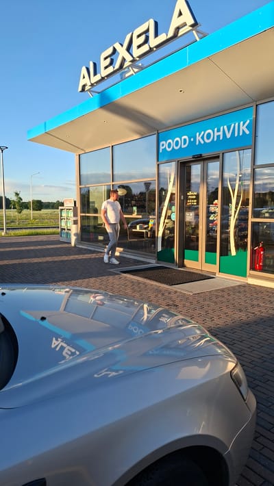 A medium shot captures an outdoor scene at a gas station with a blue awning displaying the word "ALEXELA" in white lettering. Below the awning, a blue sign reads "POOD KOHVIK". A man in a white t-shirt and grey sweatpants walks towards a glass-fronted building with two doors. He is the only person visible in the frame. The building appears to be a convenience store or cafe. In the foreground, the reflective hood of a silver car is visible, mirroring the sky and the building's signage. The ground is paved with bricks. To the left of the building, there are shelves with various products. A red fire extinguisher is mounted on the right side of the building's entrance. The environment suggests a bright, sunny day with a clear blue sky.