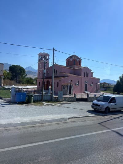 A pink Orthodox church with a bell tower and dome is visible on a clear, sunny day. The church is surrounded by a black metal fence and a paved area. Two large trash bins, one blue and one green, are located near the entrance. A white van is parked to the right of the church. In the background, there are trees, hills, and a clear blue sky. The scene appears to be in a rural or suburban area.