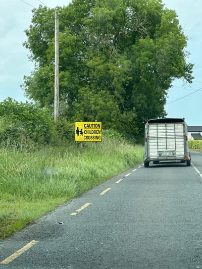 The image depicts a rural roadside scene in the vicinity of Carlow, Ireland. A bright yellow rectangular sign with black lettering and graphics is prominently displayed on the left side of the road, bearing the text "CAUTION CHILDREN CROSSING" and depicting two figures, one adult and one child, walking. The sign is surrounded by lush green grass and vegetation. A wooden utility pole stands behind the sign, supporting overhead wires. In the foreground, the asphalt road features yellow dashed lines indicating a lane divider. A trailer, presumably for livestock, is being towed by a vehicle on the right side of the road, moving away from the viewer. The trailer has a license plate that reads "01-MH-5710". The background consists of a large, dense tree with green foliage and a partially cloudy sky. The overall environment suggests a drive through the Irish countryside.