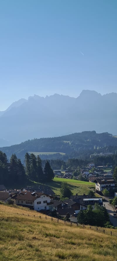 This is a high-angle, vertical, full shot of a hazy mountain landscape in Deutschnofen - Nova Ponente, Italy, during daylight. The scene is dominated by layers of mountains fading into a pale blue sky. The nearest mountains are covered in dense green forest, with a few lighter green patches of open field. Below these, a village of houses with terracotta roofs is visible. In the foreground, a sloping meadow covered in dry, golden grass is separated from the village by a simple wooden fence. A small, white contrail is visible in the upper right quadrant of the sky. No people are discernible in the image.