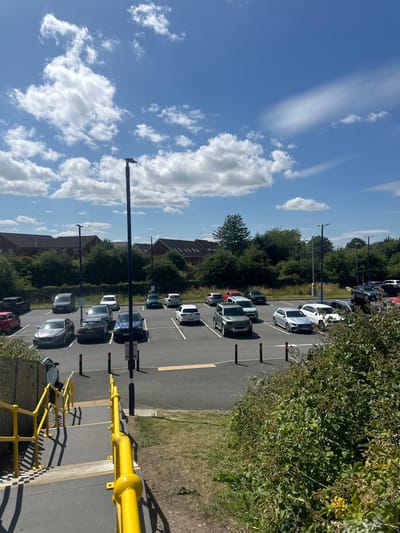 A parking lot scene under a blue sky with scattered clouds. Numerous cars are parked neatly in rows, flanked by buildings and green bushes. Tall light posts stand between the cars. A yellow railing is visible on the bottom of the frame.
