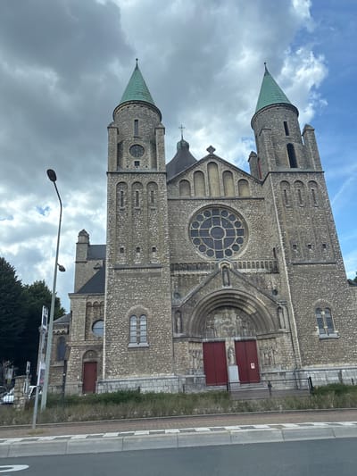 The image shows the exterior of the Saint Servatius Basilica, a Roman Catholic basilica in the city of Maastricht, Netherlands. The basilica is a large stone building with two prominent towers topped with green spires. The facade features a large rose window, a detailed tympanum above the entrance, and multiple statues. Two large red doors are visible at the main entrance. A street lamp stands to the left of the basilica, and a sidewalk and road are in the foreground. The sky is cloudy. There are no people visible in the image.