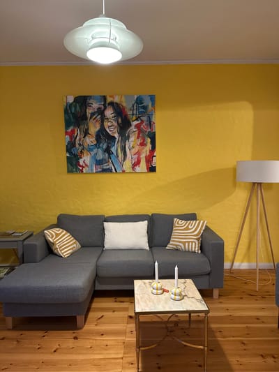 This image depicts an interior scene in Ammeldingen bei Neuerburg, Germany, likely a living room. The dominant wall color is bright yellow, creating a vibrant atmosphere. A modern, gray sectional sofa is positioned against this wall, adorned with two throw pillows: one plain white and another with a yellow and white geometric pattern. In front of the sofa, a small, marble-topped coffee table with a gold-colored metal frame holds two white candles in yellow and white striped holders, along with a red object that appears to be a lighter or small candle.

Above the sofa, a colorful abstract painting featuring figures is hung on the yellow wall. To the left of the sofa, a gray side table with magazines is visible. To the right of the sofa, a floor lamp with a white drum shade and a natural wood tripod base stands. The flooring is made of light-colored wooden planks. The ceiling features a white, multi-tiered pendant light fixture. The overall scene conveys a modern and somewhat eclectic decor style.