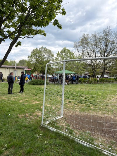 The image captures a scene at what appears to be a community park in Dulevo, Bulgaria. A soccer goal with a white net dominates the foreground. Behind the goal, people are gathered, some seated under a green canopy, suggesting a social gathering or event. Several individuals stand on the grassy field near the goal, appearing to be spectators or participants in an event. Trees with fresh spring foliage surround the open area, and a building is visible in the background, adding to the sense of a community space. The sky is overcast, and the composition of the scene is horizontal.