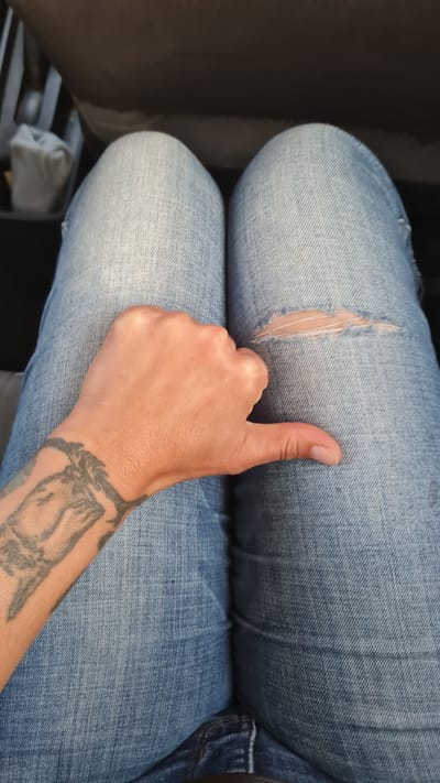 A person sits in what appears to be a car seat. They wear blue denim jeans with a rip at the upper thigh of their left leg. The person's arm, with a tattoo on the wrist, rests on their legs, giving a thumbs-up. The location is Bnei Shimon Regional Council, Israel.