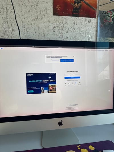 The image depicts an indoor scene in Matveevskoe, Russia, focusing on an Apple iMac desktop computer. The monitor displays a web browser open to the Zoom website (zoom.us). A cookie consent pop-up, written in Russian, is visible at the top, partially obscuring the page. Below this, a login interface in Russian prompts for an email or phone number. To the left, a promotional banner for a "Zoom PRODUCTIVITY SUMMIT EMEA" virtual event, dated "24 February 2026," is displayed, featuring small images of individuals engaged with laptops. The background consists of a light-colored, textured wall. A partially visible movie poster for "Wonder Woman," written in Russian and indicating a 2017 release date and a "12+" rating, is affixed to the wall on the right. Adjacent to this, another graphic or poster with dark, graffiti-like elements is visible. A dark cable runs vertically on the wall behind the monitor. In the foreground, a portion of a white desk is visible, along with a dark purple mousepad featuring yellow, stylized graphics. No individuals are present in the immediate scene.