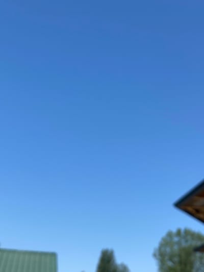 The image shows a clear blue sky. In the bottom left corner, a portion of a green roof of a building is visible. To the right of that, out-of-focus trees are present. In the upper right corner, the edge of a wooden roof or structure is visible. The scene appears to be outdoors, with no specific identifiable landmarks to pinpoint it to Arbagish, Uzbekistan.