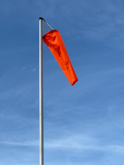 A bright orange windsock is suspended from a silver pole against a clear blue sky with wispy white clouds. The windsock, a fabric tube open at one end and closed at the other, is angled upwards and to the right, indicating the direction of the wind. It is attached to the pole by thin white ropes. The pole itself is a simple, straight, silver-colored cylinder, also oriented horizontally. The image captures a sunny day, likely daytime, given the bright lighting and clear sky. The scene is open and likely outdoors, possibly at an airport, airfield, or a location where wind direction is important for monitoring. No people are visible in the frame. The overall mood is calm and airy. There is no visible text in the image.