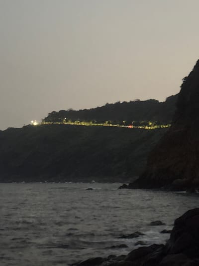 The image captures an evening or night coastal scene in Sơn Trà Ward, Vietnam. The foreground consists of dark, choppy water with scattered rocks visible along a rocky shoreline. A steep, densely forested hillside rises from the water, occupying the mid-ground and background. A continuous line of artificial yellowish-white lights delineates a road or path carved into the upper elevation of the hill, extending horizontally across the frame. Two more intensely bright white lights are present further left along this illuminated section, possibly from structures. A solitary red light, consistent with a vehicle's taillight, is observable along the illuminated path towards the right. The sky is a uniform dark grey, indicating low light conditions. No human figures are discernible.