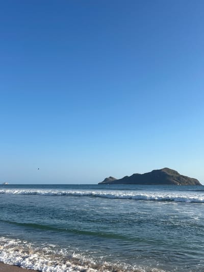 The image displays a clear coastal scene under a bright, cloudless blue sky. Approximately the upper two-thirds of the frame, particularly on the left, is filled with the expansive, azure sky, indicating clear weather.
The horizon line runs horizontally across the image. On the right side, a large, dark, rugged mountain or hill rises prominently from the ocean, its slopes extending towards the top of the frame. The base of the mountain meets the water, defining a dramatic coastline.
The ocean occupies the lower third of the image and stretches towards the right. Multiple lines of breaking waves with white crests roll towards the shore, adding dynamic texture to the water. The sea shows various shades of blue and hints of green, suggesting clarity. In the immediate foreground on the far right, white foam from receding waves is visible along the very edge of the water, likely on a sandy or rocky beach.
A small, dark, elongated speck, possibly a bird or a distant drone, is visible against the clear sky, positioned slightly above the horizon and to the left of the mountain.
The scene depicts a sunny day, likely during the mid-day hours, with calm and clear weather. No people are visible. The natural features, including the mountain meeting the sea, are characteristic of coastal regions such as Mazatlán, Mexico. There is no visible text in the image.