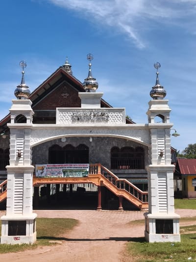 The image depicts the entrance facade of a building identified by a sign as located in Gampong Dakuta, Kecamatan Utara Batu, Kabupaten Aceh Utara, Indonesia. The central feature is a white, ornate archway with decorative molding. Two prominent, square white pillars flank the archway, each topped with a chrome-finished dome surmounted by a crescent moon and star finial. The base of the left pillar displays the number "1984," and the base of the right pillar shows "2023."

Behind the archway stands an elevated structure with grey, textured exterior walls and multiple dark-framed windows. A large, gabled roof, featuring traditional Acehnese architectural patterns in red and dark wood tones, rises above the main building. Two distinct sets of orange-brown staircases with matching railings ascend from the ground level to the elevated main entrance, which is situated behind the archway. A circular clock is visible on the wall above the main entrance.

A green and white banner, displaying images of cattle and text, is suspended centrally beneath the archway, partially obscuring the ground floor level of the building. The foreground comprises a dirt pathway leading directly to the archway, flanked by short, unkempt green grass. To the far right, a portion of a single-story building with a reddish-brown corrugated metal roof is visible. The sky is clear blue with sparse, wispy white clouds. No individuals are present in the scene.