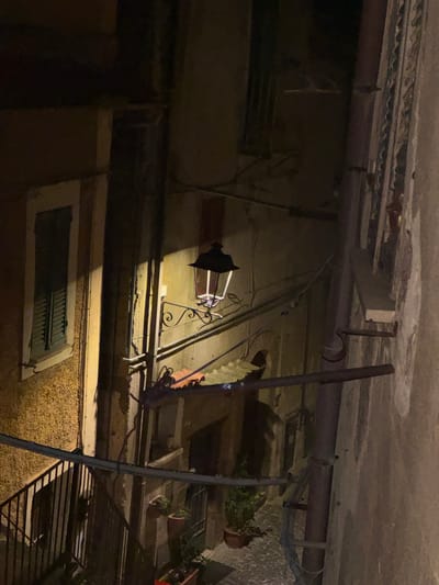An exterior shot depicts a scene in Castelnuovo di Porto, Italy, at night. The focus is on the architecture of the buildings, including a mounted lamp and a window with shutters. The image is taken from a low angle, showing the underside of architectural structures, pipes, and conduits. Potted plants are also visible on the ledge of the building. The lighting is subdued, with the mounted lamp providing a strong point of illumination.