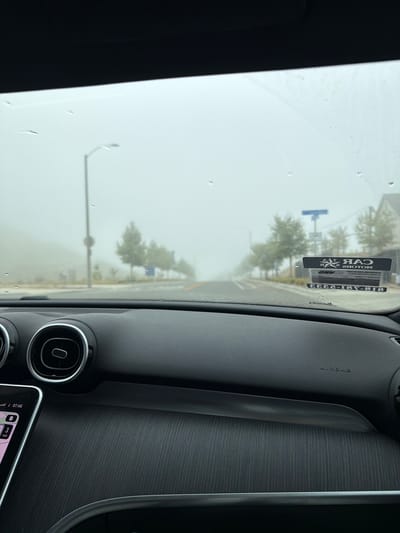 The image is taken from the interior of a car, looking out through the windshield. Heavy fog obscures the view of the road and surrounding environment. The dashboard of the car is visible in the foreground. On the windshield, there are several water droplets, suggesting it is raining or has recently rained. A license plate or sticker is attached to the windshield, which appears to be a car dealership's information. The road ahead curves to the left and is lined with trees and streetlights. The overall scene depicts a low-visibility driving condition due to thick fog. The location is identified as Santa Clarita, United States of America.
