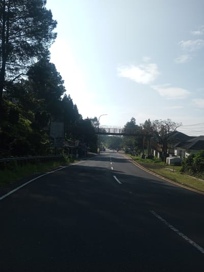 A straight, asphalt road leads into the distance beneath a pedestrian bridge. The road has white and yellow lane markings. Dense trees line the left side of the road, transitioning to buildings further along. On the right side, there is green vegetation and a building. The sky is pale blue with sparse clouds. The location is not evident to be the city of Phouviang-Gnai, Laos.