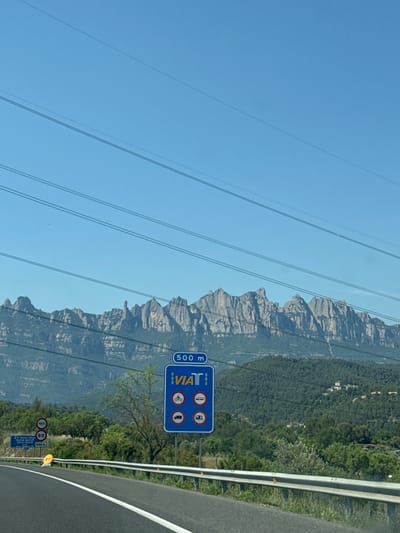 The image shows a view from a road in Castellbell i el Vilar, Spain. The scene includes a mountainous landscape with trees, power lines, road signs, and a guardrail. The road signs indicate restrictions for cyclists, buses, and motorhomes in 500 meters. Another sign indicates a speed limit. The sky is blue and clear.