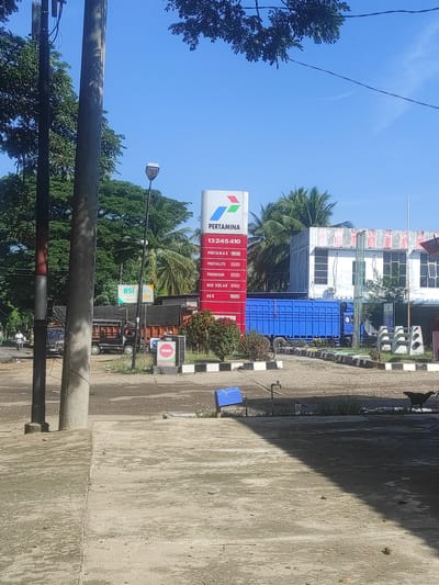 The image shows a Pertamina gas station in Kutalawah, Indonesia, on a clear day. A tall Pertamina sign with various fuel prices is prominently displayed. To the left of the sign, a lamp post stands next to a concrete pole. In the background, a white building with red and white striped trim is visible, along with palm trees and other vegetation. A blue truck with a large cargo bed is parked in front of the building. Near the gas station entrance, there is a "KELUAR" (Exit) sign. In the foreground, a concrete walkway and road are visible. The scene appears to be a typical day at a gas station with some vehicles and signs indicating commercial activity.