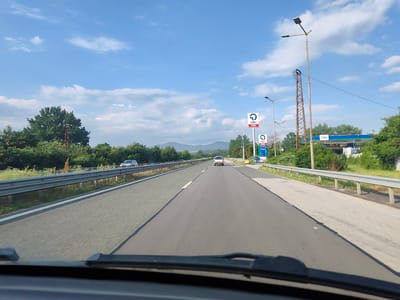 This is a point-of-view shot from a vehicle traveling on a highway, likely on the I-1 (E79) towards Osikovitsa, Bulgaria. The road is paved and divided by a white line, with a metal guardrail separating the lanes from the surrounding vegetation. On the right side of the road, there is an OMV gas station with pumps and signage. Tall streetlights and a rusty metal tower are also visible. The sky is blue with scattered white clouds, and a range of hills can be seen in the distance. A few vehicles are present on the highway, including a silver minivan and a white SUV. The image appears to have been taken during daytime, under clear weather conditions.