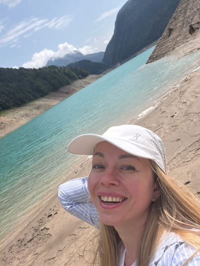 A blonde woman wearing a white baseball cap and a blue and white striped shirt is taking a selfie. She is smiling and looking at the camera. The woman is standing on a sandy embankment next to a large, turquoise body of water. In the background, there are mountains covered in lush green trees. The sky is blue with scattered white clouds. The image appears to be taken during the daytime, under bright sunlight.