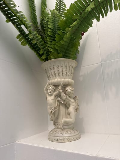 A close-up shot captures a decorative cream-colored vase filled with artificial fern fronds. The vase is adorned with reliefs of cherubs, appearing to hold up the rim. The vase and ferns are positioned on a white shelf against a white tiled wall. The scene is well-lit, creating subtle shadows from the fronds on the wall. The specific location in Tha Chana, Thailand is not discernible from the image itself.