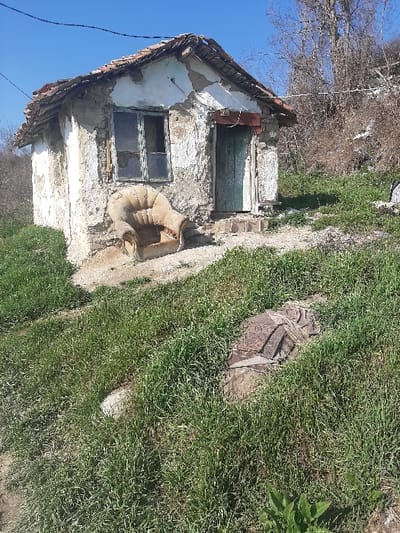 The scene is an exterior view of a dilapidated, single-story structure in what is reported to be the city of Ivanski, Bulgaria. The building, constructed from stone or mud covered in peeling, whitewashed stucco, features a damaged tile roof with exposed wooden beams. A multi-paned window with broken glass and a weathered frame is visible on the left side of the facade. To the right, a weathered, faded green door is set into the wall, accessed by two small, rough steps. Directly in front of the house and slightly to the left, a heavily worn, tan or beige upholstered armchair with a high back rests on the bare ground. The immediate foreground and surrounding area are characterized by unkempt green grass mixed with patches of exposed dirt. Further down the grassy slope, to the right and partially obscured by vegetation, lies an indeterminate object, possibly a weathered tire or fabric, showing patterned sections. In the background, beyond the house and rising terrain, are bare trees and dry brush against a clear blue sky. No individuals, direct actions, or interactions are visible. Overhead, thin wires stretch across the frame, connecting to an unseen point.
