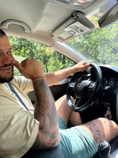 This image captures a low-angle, sideways view inside a car in Dorkovo, Bulgaria, during what appears to be a sunny day. A man is in the driver's seat, his face visible with dark hair and a beard, looking towards the camera. He is wearing a light-colored t-shirt and green shorts.
His left hand is on the black steering wheel, which features integrated controls. His right hand is raised, with his thumb touching his nose. Both his forearms and left leg are heavily tattooed. A prominent tattoo on his right wrist reads "АЛИНОЧКА МОЯ НАВСЕГДА," which translates from Russian to "Alinochka my forever." His left leg displays several cartoon-like figures.
The car's interior details include a dashboard with visible gauges, one appearing to show "185" (possibly speed in kph), and keys hanging from the ignition. The gear shifter is marked with "DSG". A sun visor is down, with a small photograph tucked into it. A rearview mirror and a passenger grab handle are also visible. Through the car windows, abundant green foliage is seen, indicating an outdoor, natural environment.