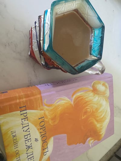 A close-up, overhead shot shows a faceted, translucent blue glass mug filled with light brown liquid, likely coffee, resting on a light-colored, marbled surface. The mug is positioned over an open book, partially obscuring it. The book has a predominantly pink cover with an illustration of a silhouette of a blonde woman with her hair in a bun. Russian text is visible on the book cover. To the left of the mug, a stack of what appears to be candy wrappers, in red, white, and blue, is stacked. The overall lighting is bright, suggesting an indoor setting with overhead illumination. There are no people or animals visible in the frame. The city of Lozhki, Russia, is not depicted in this image.