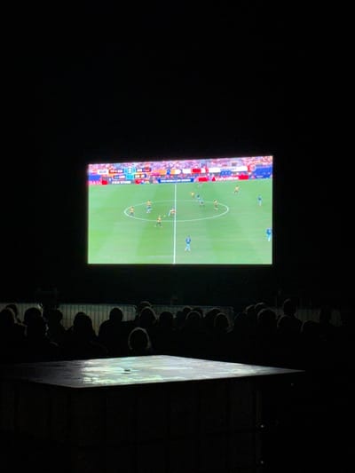 This image depicts an outdoor screening of a soccer match in Langeoog, Germany. The main subject is a large screen displaying the game in progress. The score displayed on the screen indicates a match between Ecuador (ECU) and Germany (GER), with Ecuador leading 2-1. The players are visible on the green field, and a crowd of silhouetted figures is gathered in the foreground, watching the screen. The environment is dark, suggesting it is nighttime, and a portion of a table or platform is illuminated in the lower foreground by light from the screen.