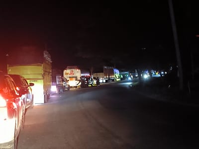 The image depicts a traffic jam at night in Gampongrayadagang, Indonesia. A line of vehicles, including cars and trucks, are stopped on the road. Some trucks have the number "35" displayed, and one has signage indicating it is carrying flammable liquids. A person on a motorbike is navigating between the vehicles. The surrounding environment is dark, with trees visible on the side of the road. The scene is illuminated by vehicle headlights and taillights.