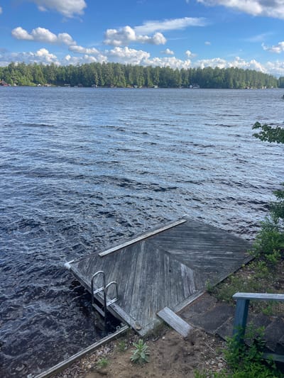 The image depicts a waterfront scene at a lake, possibly in Brantingham, United States. A weathered wooden dock extends into dark, rippling water. A silver ladder is attached to the dock, with its lower rungs submerged. The shore adjacent to the dock is composed of sand, small stones, and sparse vegetation. Further back, a blue wooden railing is visible, leading up some steps. In the background, a dense line of green trees forms the far shoreline, with several small buildings nestled among them. The sky above is a vibrant blue, dotted with white, fluffy clouds. The overall impression is one of a tranquil, natural setting.