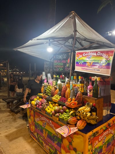 A vibrant fruit and drink stand is displayed with various fruits and liquor bottles arranged on shelves. The stand is decorated with colorful signs that read "Faz Gostoso Drinks" and promotes a drink special. An employee stands behind the stand, attending to customers. The stand is covered by a large umbrella, and the surrounding environment suggests a nighttime outdoor setting with other people in the background.