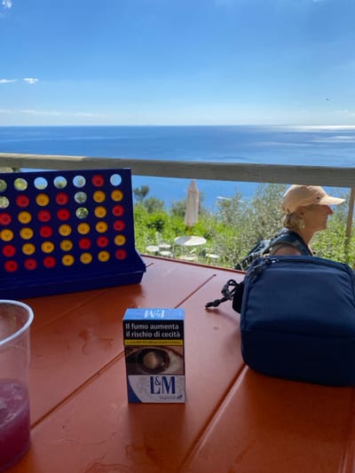 The image captures an outdoor scene in Corniglia, Italy, under a clear blue sky with minimal cloud cover, overlooking the Mediterranean Sea. In the immediate foreground, an orange-brown outdoor table displays several items. On the far left, a clear plastic cup contains a reddish-purple liquid with ice. To its right, a blue "Connect Four" game board stands upright, populated with red and yellow tokens. Centrally located is a blue and white pack of L&M cigarettes, oriented vertically, featuring the Italian health warning "IL FUMO AUMENTA IL RISCHIO DI CECITA'" (Smoking increases the risk of blindness) and an image of an eye. To the right of the cigarette pack, a small, dark blue zippered bag with a braided cord handle rests on the table. Water rings are visible on the table surface.
To the right, partially visible from the chest up and in profile, is an adult individual with light-colored hair, wearing a beige baseball cap and a dark top, facing right and looking out towards the sea. A visible earring adorns their ear. Behind the table, a horizontal wooden railing separates the foreground from the distant view. Beyond the railing, a green, terraced hillside populated with olive trees descends towards the coastline. White outdoor tables and umbrellas are visible on the hillside, suggesting a seating area. The expansive, deep blue Mediterranean Sea stretches to the horizon, reflecting sunlight on its surface.