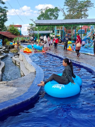 A woman in a blue and white patterned shirt and black pants sits on a blue inflatable tube in a lazy river. The river is lined with gray concrete and a blue and white tiled border. The water is blue and clear. The river is part of a water park, with a metal roofed structure overhead. There are a number of other people in the water park, including children and adults, playing and swimming. Some of the people are wearing life jackets, while others are wearing swimsuits. The day is bright, sunny, and cloudless. The location is in Kecamatan Tulung, Indonesia (latitude: -7.592, longitude: 110.65).