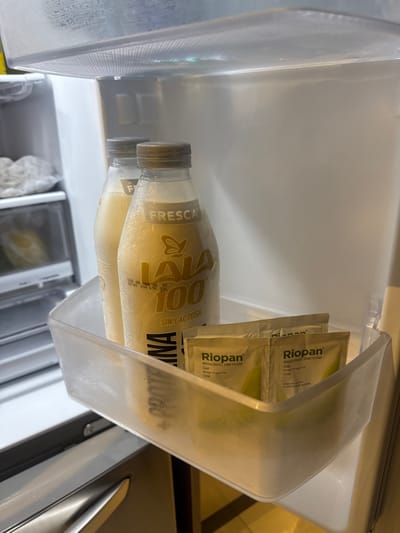 A fridge shelf contains two bottles of "LALA 100" lactose-free milk, labeled "FRESCA." Two packets of "Riopan" Magaldrato Dimeticona oral gel are visible behind the bottles. The shelf itself is plastic and appears frosted or slightly dirty. Text on the shelf is partially obscured but reveals the word "PROTEANA."