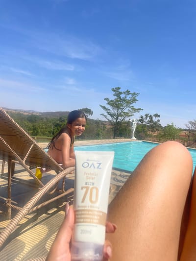 The image captures an outdoor scene under a clear blue sky, likely within the Pirenópolis, Brazil region, characterized by its rolling green landscape. In the immediate foreground, a person's bare legs are visible on the right, while a hand on the left holds a white tube of "OAZ Protetor Solar FPS 70" sunscreen (200ml), bearing Portuguese text indicating high UVA/UVB protection, water resistance, and application for body and face. In the midground, a young female child, approximately 7-10 years old, with dark wet hair and wearing a dark bikini top, is seated on a light brown wicker lounge chair to the left. She smiles directly at the camera. Adjacent to her and extending into the background is a light blue swimming pool with decorative tile edging. The background consists of verdant hills and scattered trees. A light-colored, classical-style statue of a male figure stands near the far edge of the pool, partially obscured by foliage. The scene is brightly illuminated by direct sunlight.