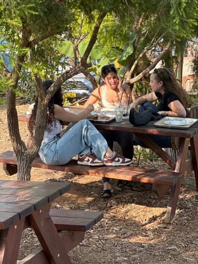 Three females are seated around a wooden picnic table outdoors in Kfar Monash, Israel, under a canopy of green trees. The ground is covered with wood chips.

The female on the left, partially obscured by a tree trunk, has long, dark curly hair and wears a white sleeveless top, light blue denim jeans, and brown and white athletic shoes. She is positioned with her right arm on the table, appearing to look down at the items in front of her.

The female in the middle has dark hair pulled back with sunglasses on her head, a white sleeveless top, dark pants, and black flat sandals. She is actively speaking, smiling, and gesturing with her left hand near her face, looking towards the female on her right.

The female on the right has long, wavy brown hair and wears a dark top and dark pants. She is looking towards the middle female, appearing to be engaged in conversation. A black handbag rests on the table next to her.

On the table are multiple plastic and paper cups with straws, metal trays containing food remnants, and a white plate. Sunlight filters through the tree leaves, creating dappled light. In the background, a light-colored vehicle with a partially visible Israeli license plate (G-68-XX) is discernible through the foliage, along with indistinct building structures. A second wooden picnic table is partially visible in the foreground.